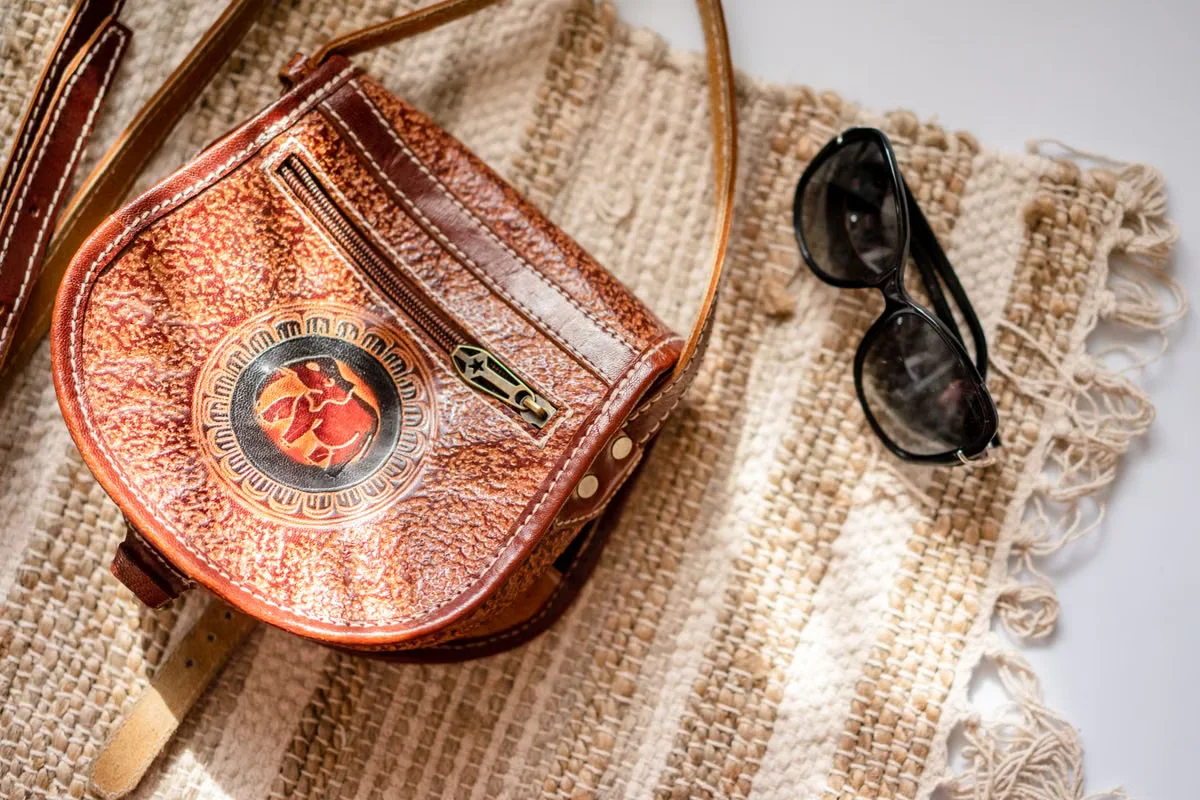 Brown hand-tooled leather crossbody bag laid flat beside sunglasses on a woven textile surface — the bag's compact proportions illustrate a typical mini crossbody format sized for phone, cards and keys