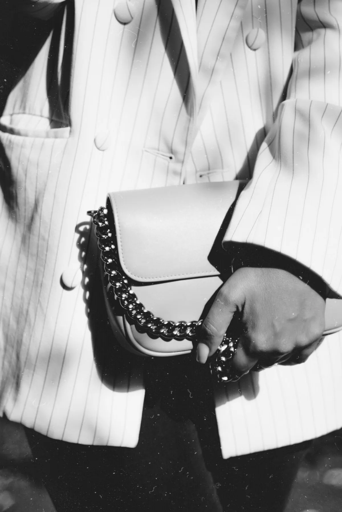 Black and white photo of a woman holding a small chain crossbody bag against a white pinstripe blazer — illustrating how crossbody bag proportions relate to the human body, which is the fundamental reference point for all crossbody dimension decisions