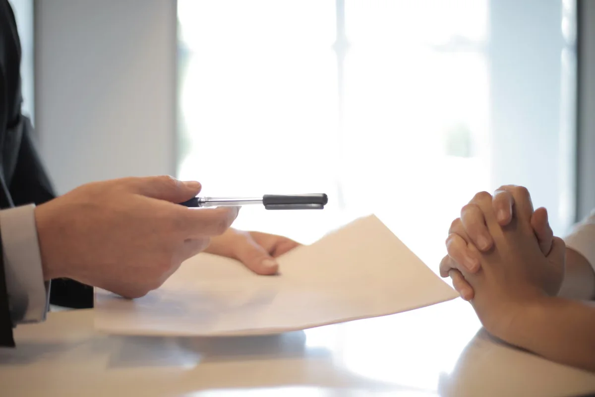 A business professional presenting a pen and contract to a counterpart across a table — successful leather bag factory price negotiation ends with a written agreement that both parties are committed to fulfilling