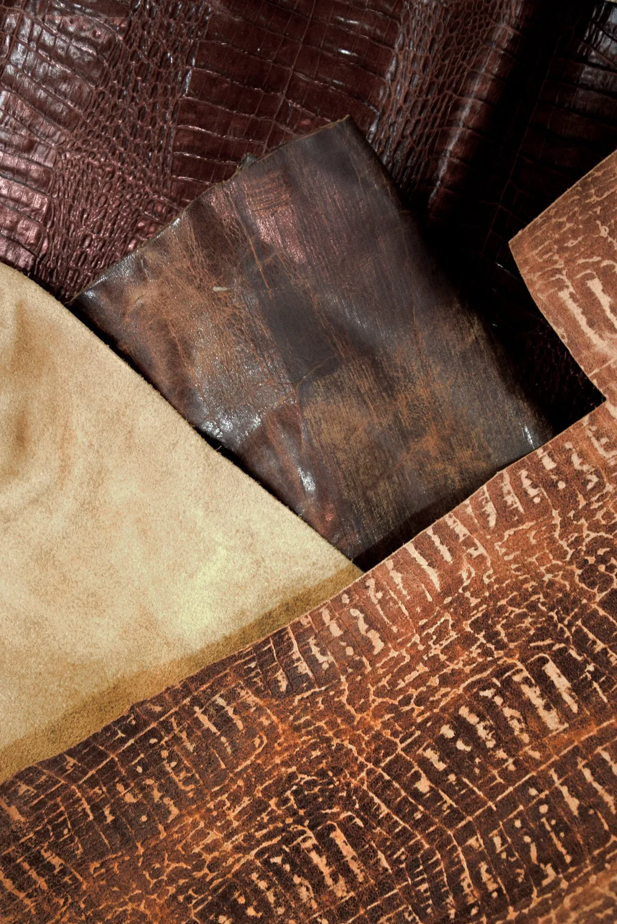 Multiple leather hide samples layered together showing contrasting textures — crocodile-embossed, smooth full-grain, and suede — illustrating the wide range of leather grades and finishes available in wholesale handbag sourcing