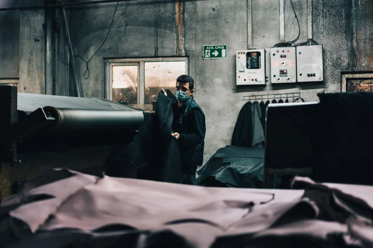 A leather tannery worker handling a large roll of processed dark leather in an industrial factory setting — quality and scale of raw material sourcing directly determines factory MOQ thresholds